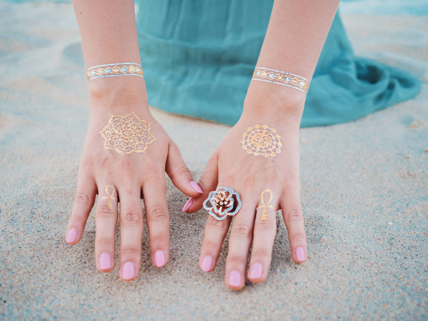 Silver and golden flash tattoo on female hands over sea or ocean background. close up hands with boho gypsy gold accessories. Woman touching sand on the beach at summer - Photo, Image