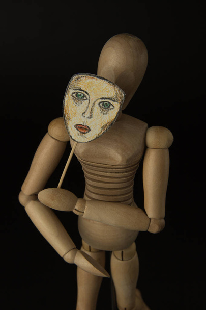 Muñeca de madera en bisagras sostiene una máscara en las manos y cubre su cara sobre un fondo negro
 - Foto, imagen