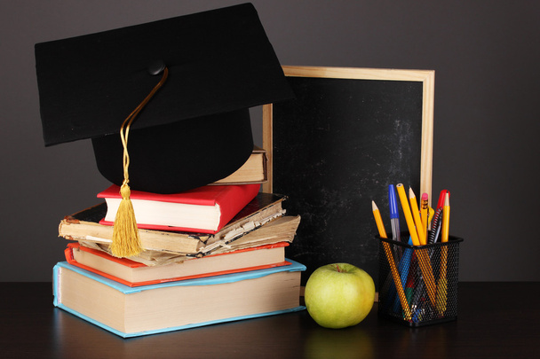 Books and magister cap against school board on wooden table on grey background - Photo, Image