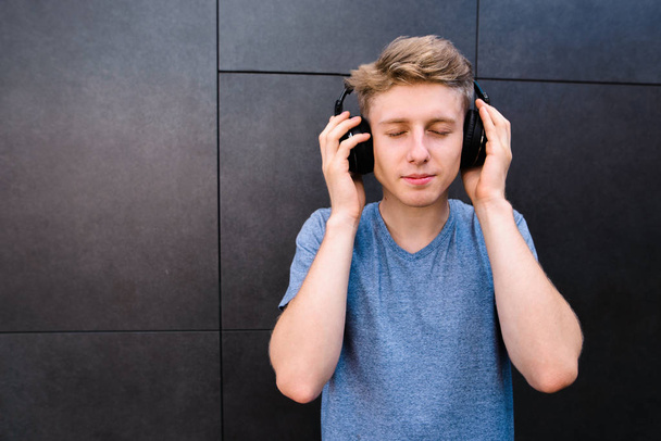 Όμορφος νεαρός άνδρας με τα μάτια του κλειστά να ακούτε μουσική στα ακουστικά του στο φόντο της ένα γκρίζο τοίχο. Ο έφηβος απολαμβάνει να ακούτε μουσική - Φωτογραφία, εικόνα