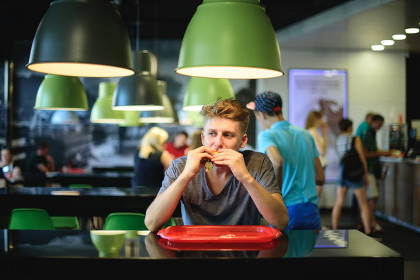 The teenager is sitting in a fast-food restaurant, eating a large, beautiful burger and looking away. - Photo, Image