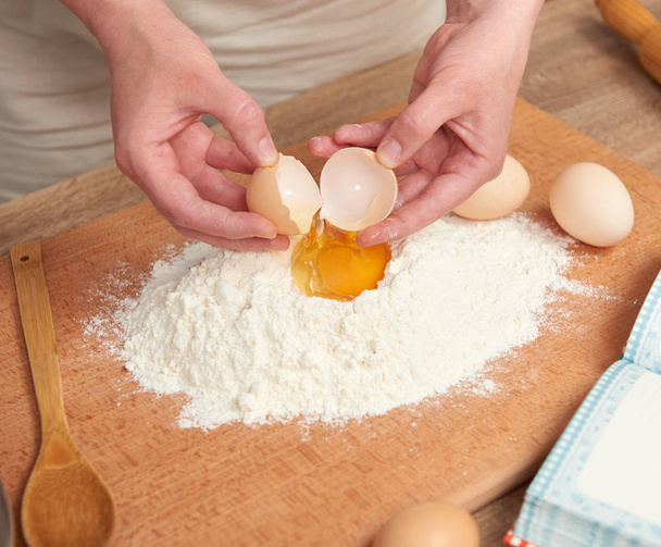Frau bereitet Mehl zum Backen auf einem hölzernen Hintergrund zu. Rohkost und Küchenutensilien. Hände und Lebensmittel in Nahaufnahme. Ein Ei brechen, Teig kochen. - Foto, Bild