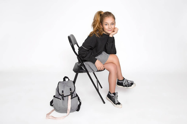Linda niña vestida con ropa de escuela y mochila escolar sentada sobre fondo blanco
 - Foto, imagen