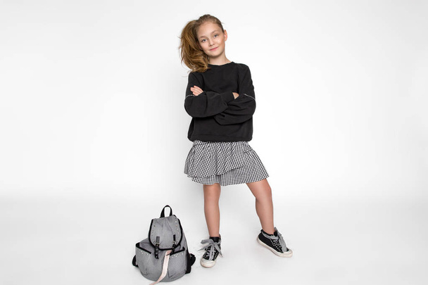 Cute little girl dressed in school clothes and school backpack sitting on white background - Foto, imagen
