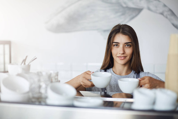 Female barista holding coffee and tea cups for cappuccino ready to put them onto an espresso machine to warm them up - Foto, Imagem
