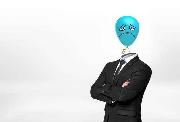 A businessman on white background stands with crossed hands and a blue sad face balloon instead of his head. - Фото, изображение