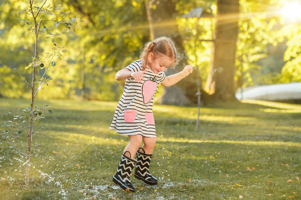 The cute little blond girl in rubber boots playing with water splashes on the field in summer - Zdjęcie, obraz