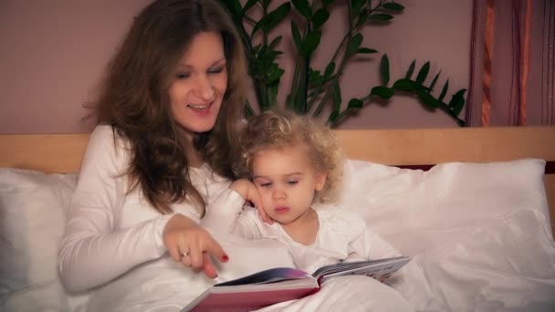 pretty young mother reading book to her cute toddler daughter sitting in bed - Felvétel, videó