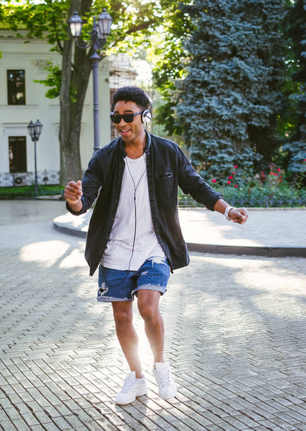 Portrait of young stylish hipster black man in white headphones and sun glasses dancing outdoor in city centre, having some fun - Photo, Image