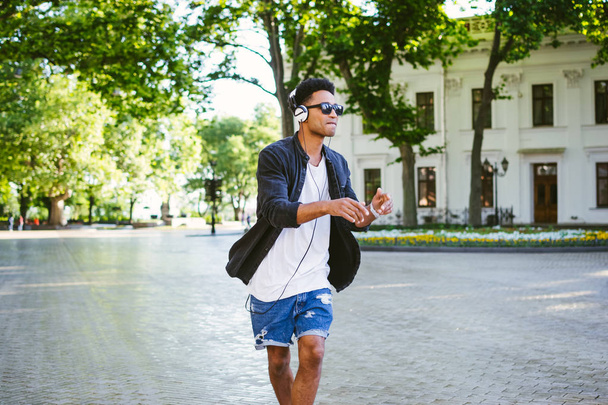 Portrait of young stylish hipster black man in white headphones and sun glasses dancing outdoor in city centre, having some fun - Фото, зображення