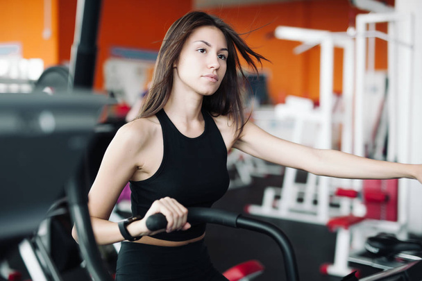 Young beautiful white girl in a sports suit is engaged on a stationary bike in the fitness club. - Foto, imagen