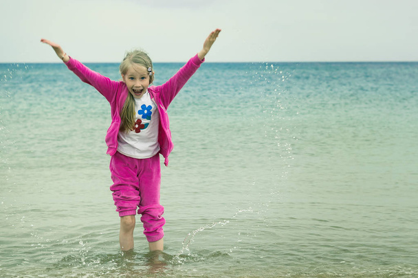Cheerful girl in a warm suit plays with water in a cold sea. - Photo, Image