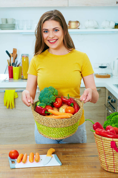smiling woman standing in kitchen holding basket with vegatables - Фото, зображення