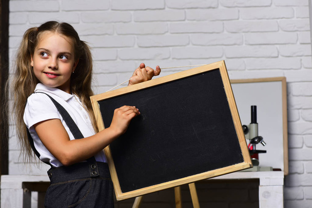 Schoolgirl with cunning face holds small blackboard - Foto, Imagen