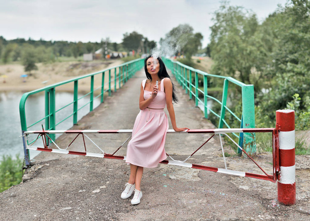 mujer vapeo dispositivo de vapor mod en la naturaleza. concepto de vapeo. mujer usando en vestido rosa y zapatos blancos
 - Foto, Imagen