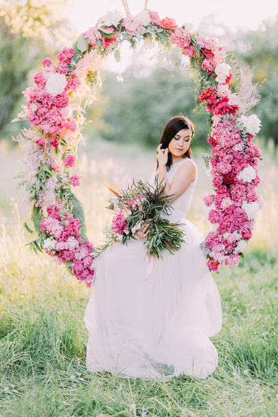 The portrait of the cute bride holding the bouquet of flowers and sitting in the wedding peonies arch in the sunny field. - Foto, Imagen