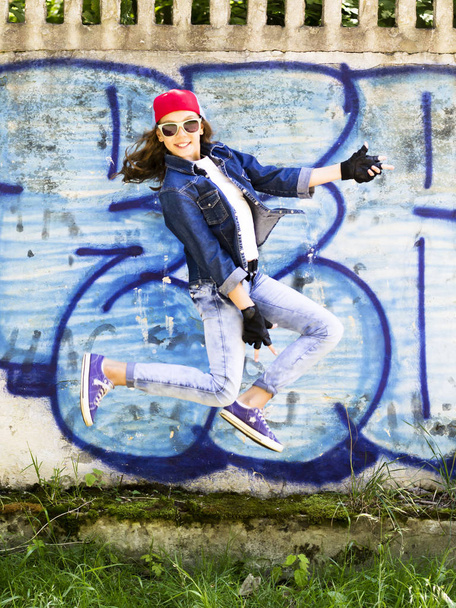 Roztomilá mladá blondýna dospívající dívka v baseballovou čepici a jeans košile, skákání na pozadí kamenné zdi. Hip hop, - Fotografie, Obrázek