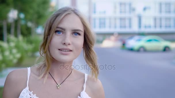 Young and unbelievably beautiful blonde woman with sky-blue eyes in white dress walks down the alley and nicely smile while combing her hair. - Felvétel, videó