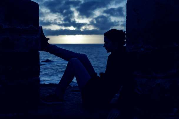 Silhueta de uma jovem mulher contemplando sentado em uma parede sobre
 - Foto, Imagem