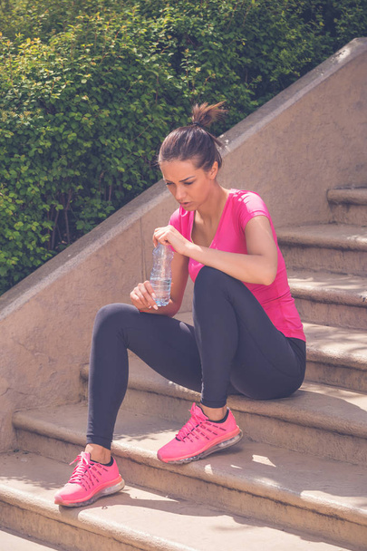 Female jogger sitting on stairs and holding bottle of water  - Фото, изображение
