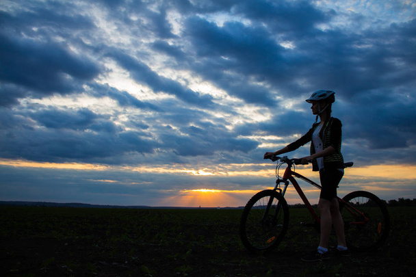 Sylwetka młodej kobiety z rowerem w polu o zachód słońca niebo z chmurami dramatyczne - Zdjęcie, obraz