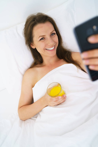 Femme mignonne vérifie le smartphone et boit du jus d'orange au lit
  - Photo, image