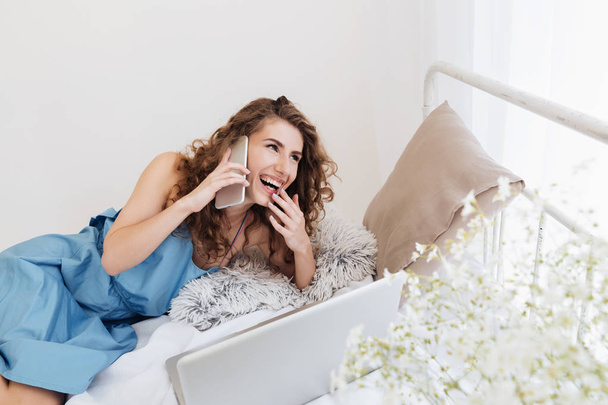 Woman sitting indoors on bed talking by phone. Looking aside. - Foto, imagen