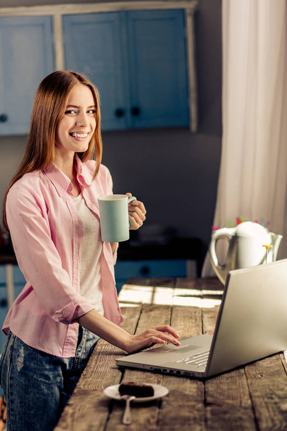 Happy young girl working on laptop while drinking coffee or tea. - Foto, Imagen