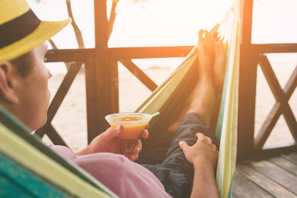 Handsome young man holding an orange cocktail and lying in a hammock at a sunny beach by an ocean - 写真・画像