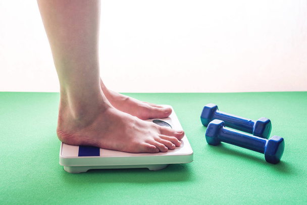 Female feet standing on mechanical scales, dumbbells. Concept of sports training, slimming and weight loss - Photo, Image