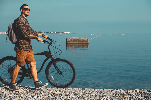 Young Man Cyclist In Sunglasses With Bicycle Walking On Coast And Enjoying View Of Sea. Holiday Travel Activity Concept - Zdjęcie, obraz