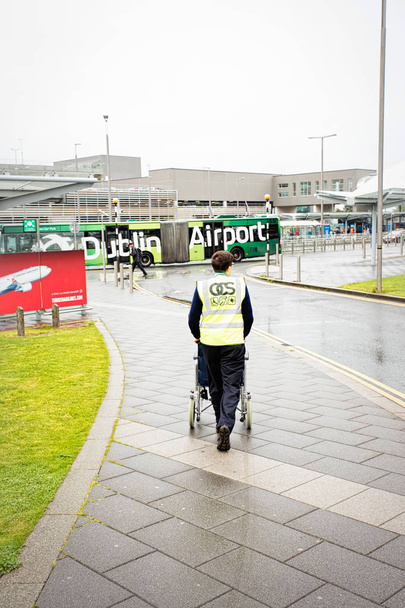 Mitarbeiter des Dubliner Flughafens schieben Rollstuhl für behinderten Passagier zum Flughafengebäude an einem regnerischen Sommertag, Dubliner Flughafen, 14. August 2017 - Foto, Bild