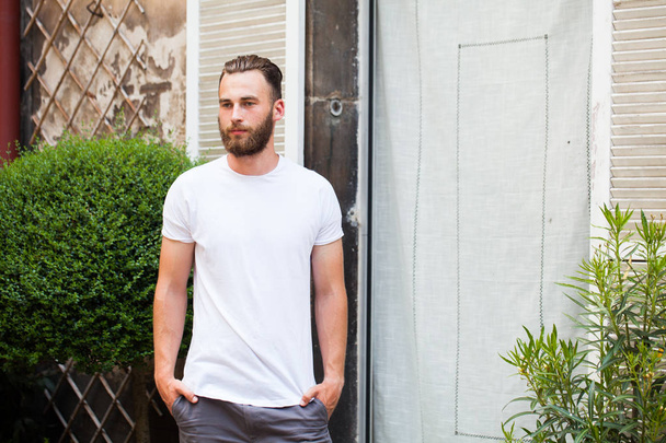Hipster handsome male model with beard wearing white blank t-shirt with space for your logo or design in casual urban style - Fotó, kép
