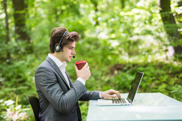Straně portrét podnikatel s mikrofonem sedí na kancelářský stůl s přenosným počítačem a šálek kávy v call centru v zeleným lesoparkem. Obchodní koncept. - Fotografie, Obrázek