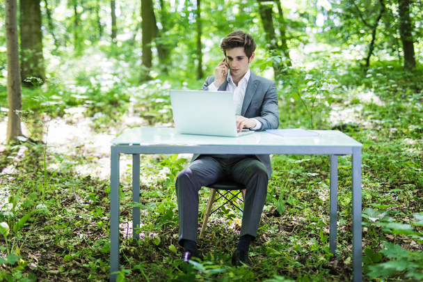 Νέοι επαγγελματίες όμορφος άνθρωπος κάθεται στο γραφείο στο καταπράσινο πάρκο ή δάσος και εργασία στο φορητό υπολογιστή, ενώ μιλάμε στο τηλέφωνο με πελάτης. Επιχειρηματική ιδέα. - Φωτογραφία, εικόνα