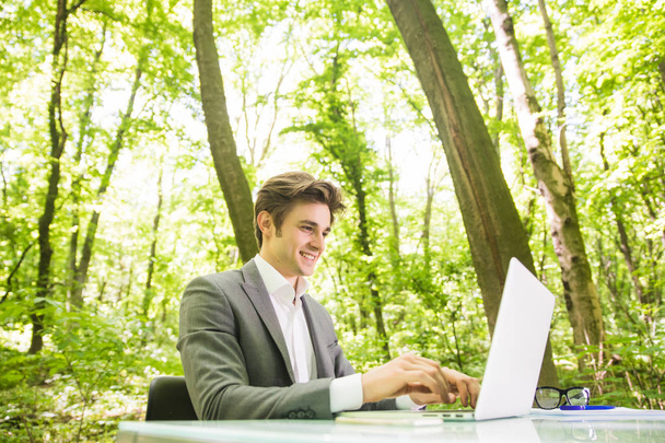 Portret stronie młody przystojny działalności człowieka w garniturze działa na laptopie w tabeli office w zielonym parku leśnego. Koncepcja biznesowa. - Zdjęcie, obraz