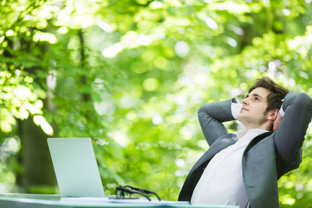 Portrét mladých pohledný obchodní muž v obleku relaxovat v notebooku u stolu office ruce nad hlavou v zelené forest park. Obchodní koncept. - Fotografie, Obrázek