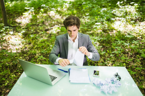 Joven hombre de negocios guapo en la oficina de mesa de trabajo con ordenador portátil en papeles de trabajo de lágrimas de bosque verde. Concepto empresarial
 - Foto, imagen