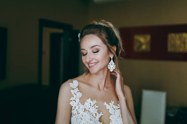 Blonde bride puts on earrings in the morning in the room - Photo, Image
