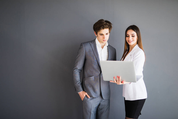 Beautiful young business woman and handsome businessman in formal suits are using a laptop, talking and smiling, standing before gray wall - 写真・画像