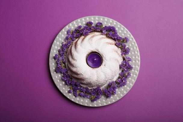 Top view of a ring cake sprinkled with powdered sugar, with a violet candle in a center on a bright violet background. - Фото, изображение
