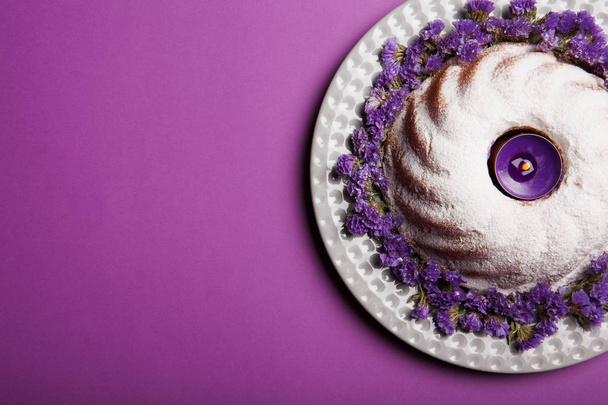 A plate with round cake sprinkled with powdered sugar and a candle in the center on a bright violet background, top view. - Foto, afbeelding
