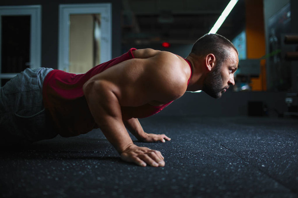 Side view of young handsome man in sportswear doing push-up at gym. Handsome man with a muscular body on a blurred background. - Foto, Imagem