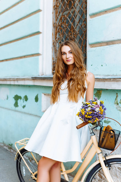 Κοπέλα με λευκό φόρεμα με ποδήλατο και λουλούδια κοντά μπλε τοίχο - Φωτογραφία, εικόνα