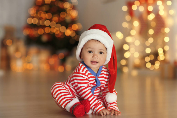 Cute little baby with Santa hat on floor and blurred Christmas lights on background - Foto, imagen