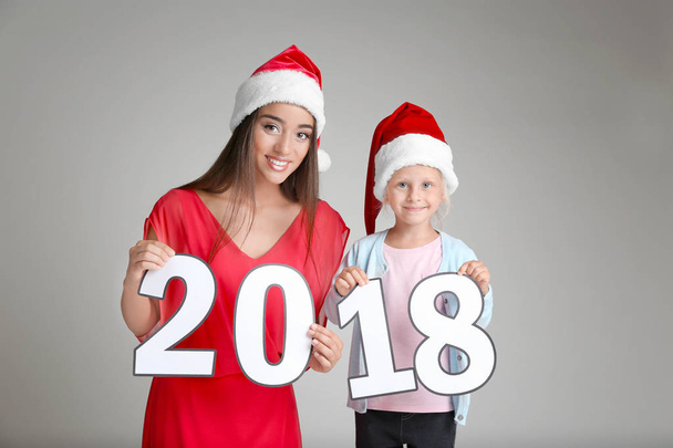 Young woman and cute little girl in Santa hats with paper figures 2018 on light background. Christmas celebration concept - 写真・画像