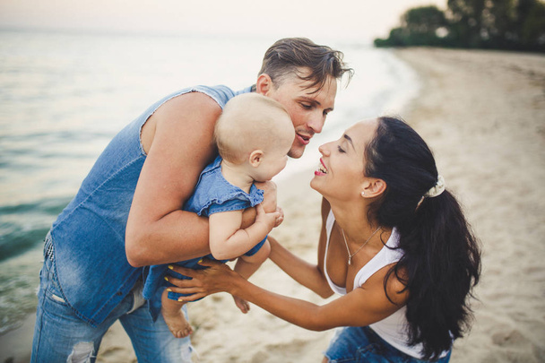 Üç genç, güzel bir aile. Anne, baba ve kızı babası oyunumu kollarında sevinirler, sahilde kum plajındaki yaz aylarında gülümse. Junky giysi ve beyaz gömlek giymiş - Fotoğraf, Görsel