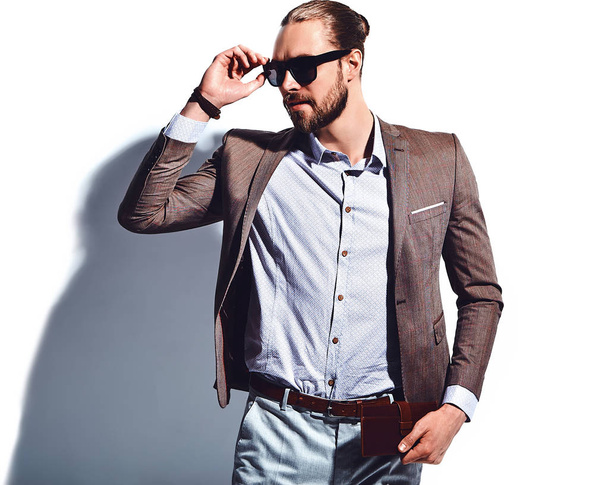 portrait of handsome fashion stylish hipster businessman model dressed in elegant brown suit in sunglasses posing near white wall in studio. Pulls out or put his leather wallet into jacket pocket - Foto, immagini