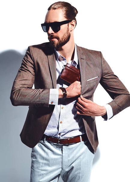 portrait of handsome fashion stylish hipster businessman model dressed in elegant brown suit in sunglasses posing near white wall in studio. Pulls out or put his leather wallet into jacket pocket - Photo, image
