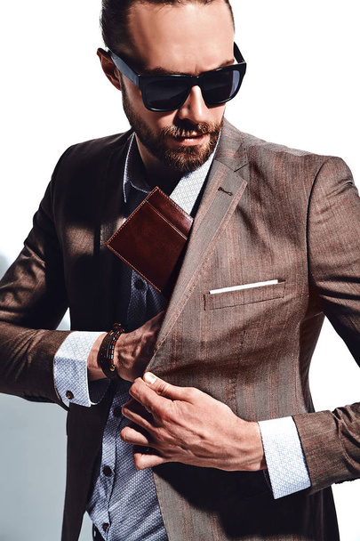 portrait of handsome fashion stylish hipster businessman model dressed in elegant brown suit in sunglasses posing near white wall in studio. Pulls out or put his leather wallet into jacket pocket - Fotoğraf, Görsel